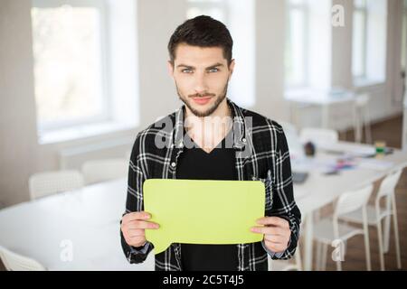 Young man in black short thoughtfully looking in camera while holding yellow paper shape of message in hands at work in modern empty office Stock Photo
