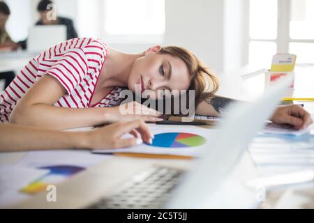 Young tired woman in striped T-shirt lying on desk dreamily looking aside while spending time in modern office Stock Photo