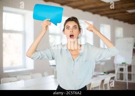 Young woman in blouse amazedly looking in camera holding blue paper message icon in hand near head while spending time in office Stock Photo