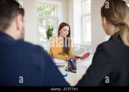 Young businesswoman in blouse dreamily working with diagrams while spending time with colleagues in modern office Stock Photo