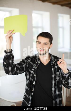 Young man with beard dreamily looking in canera with green paper message icon in hand near head while showing thumb up gesture in modern office Stock Photo
