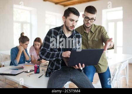 Young guys in eyeglasses and shirts with beard thoughtfully using laptop while working together in office with colleagues on background Stock Photo