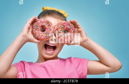 Funny little girl on background of bright blue wall. Beautiful child is having fun with donut. Yellow, pink and turquoise colors. Stock Photo