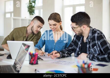 Young creative people with laptop working on project together. Group of joyful workers spending time in modern office Stock Photo