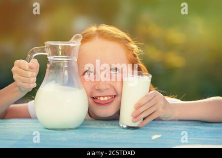 Milk is fresh from the cow - a beautiful girl with dairy products. Girl holds glass with fresh milk. Happy redhead child in summer. Closeup of little Stock Photo