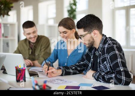Young man in shirt and eyeglasses thoughtfully working in office with colleagues on background Stock Photo