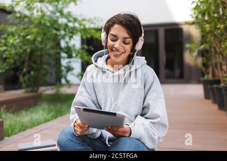 Pretty smiling lady sitting in headphones and tablet in hands while happily spending time in courtyard of university Stock Photo