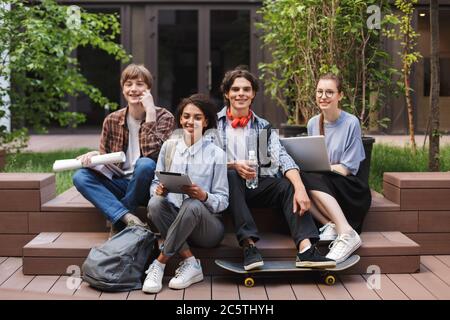Group of cool smiling students sitting and happily looking in camera while spending time together in courtyard of university Stock Photo