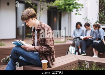 Young thoughtful man sitting on bench with coffee and writing in notebook while spending time in courtyard of university with students on background Stock Photo
