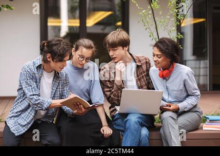 Photo of students sitting on bench with laptop and book while spending time in courtyard of university Stock Photo