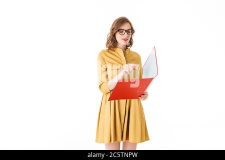 Portrait of pretty thoughtful lady in eyeglasses and yellow dress standing with red folder in hand on white background isolated Stock Photo