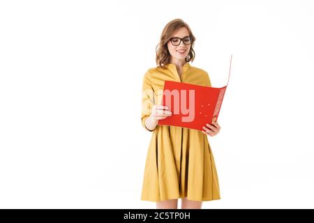 Portrait of pretty smiling lady in eyeglasses and yellow dress standing with red open folder in hand on white background isolated Stock Photo