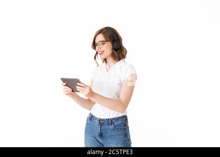 Portrait of beautiful smiling lady in eyeglasses and black headphones standing and happily looking in tablet on white background isolated Stock Photo
