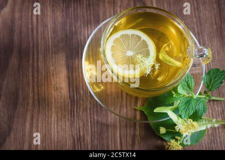 A cup of medicinal, lime tea, standing on a wooden table, in the rays of sunlight, there are flowers of chamomile lime trees around. Stock Photo