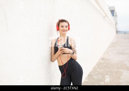 Young smiling woman in dark gray sporty top and leggings in red headphones joyfully looking aside leaning on wall while holding cellphone in hands Stock Photo