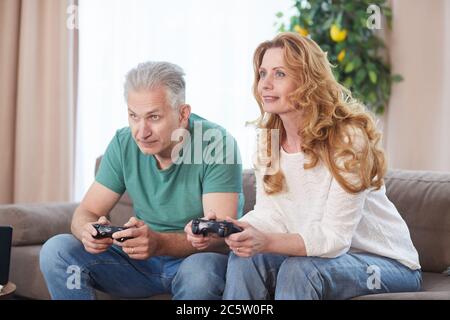 Portrait of modern mature couple playing video games via console and holding controllers while enjoying time at home Stock Photo