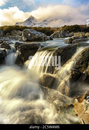 Flowing Waterfall In Sligachan With Morning Sunlight And Scottish Snowcapped Mountains In Background. Isle Of Skye, UK. Stock Photo