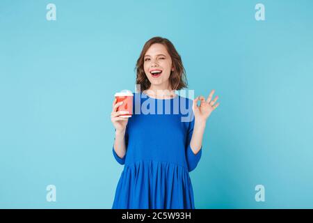 Young beautiful lady in dress standing with cup of coffee to go on over blue background Stock Photo