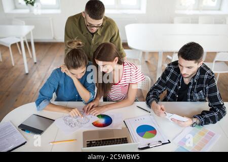 Group of thoughtful men and women working with sketches and laptop while spending time at work. Young creative people discussing new project together Stock Photo