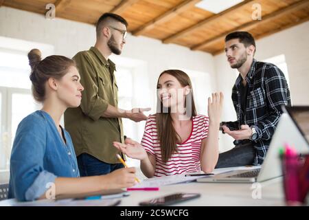 Two smiling girls happily talking while spending time in modern office with colleagues on background. Group of creative people working together Stock Photo