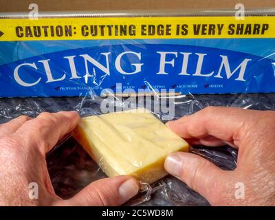 Man’s hands wrapping cling film around a piece of cheese. The tear-off roll behind, shows a warning as to the serrated cutting edge being very sharp. Stock Photo