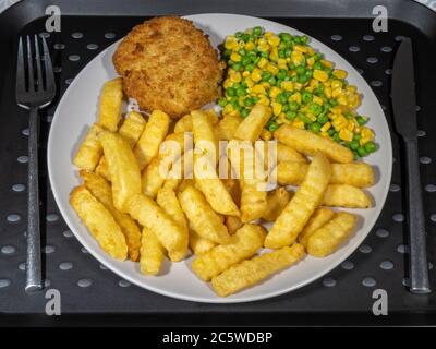 Closeup POV overhead shot of a knife and fork next to the dinner plate of hot crispy baked fishcake and chips / fries, with peas and sweetcorn. Stock Photo