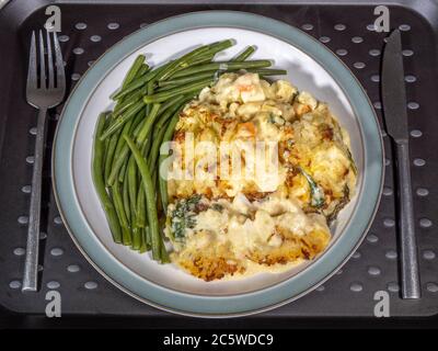 Closeup POV overhead shot of a knife and fork next to the dinner plate of hot baked fish and vegetable pie, surrounded by green beans. Stock Photo