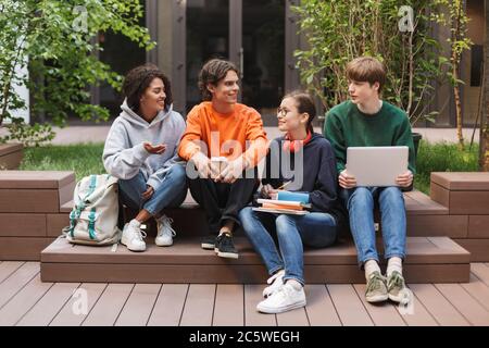 Group of cool smiling students sitting and happily looking at each other while spending time together in courtyard of university Stock Photo