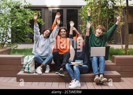 Group of joyful students sitting and happily looking in camera while raising their hands up in courtyard of university Stock Photo