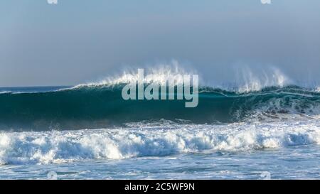 Ocean wave feathering upright wall of blue water flaring and crashing on shallow reef sandbars closeup panoramic photo. Stock Photo