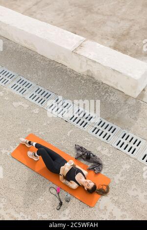 Young nice woman in sporty top and leggings with earphones holding cellphone in hands while dreamily lying on orange yoga mat with jump rope and sport Stock Photo