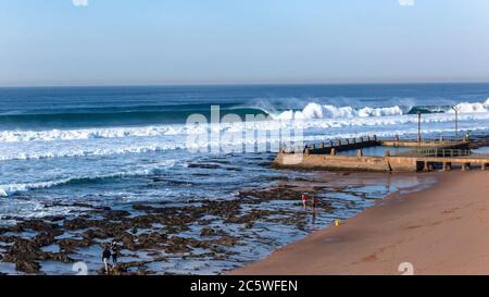 Beach tidal swimming pool on reef coastline with ocean wave walls of blue water crashing with distant public on shoreline. Stock Photo