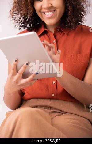 Happy young elegant woman with curly hair and toothy smile touching screen of tablet while scrolling through posts in social networks Stock Photo