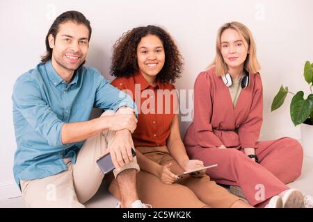 Three young cheerful friends in casualwear sitting by wall in studio and using modern mobile gadgets for online communication Stock Photo