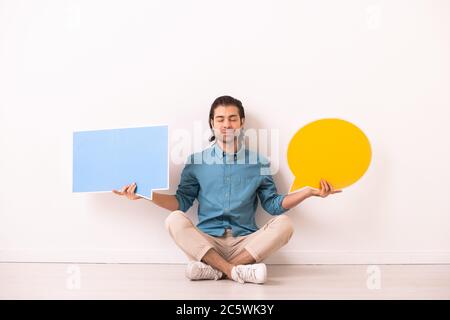 Young serene man in denim shirt and white pants holding two blank paper speech bubbles while sitting on the floor with his legs crossed Stock Photo