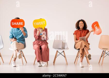Three young casual friends with mobile gadgets holding multi-color paper speech bubbles while communicating in social networks Stock Photo