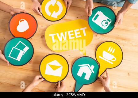 Overview of millennials hands holding chosen paper speech bubbles with social networks icons around yellow one on wooden table Stock Photo