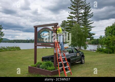 A worker standing on a ladder installing a sign for the Speculator Public Park on Lake Pleasant in the Adirondack Mountains, NY USA Stock Photo