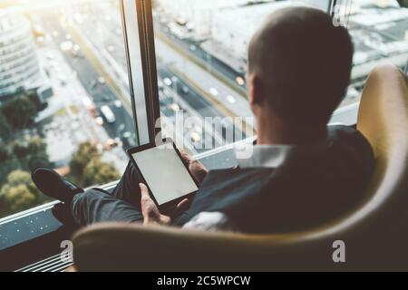 View from behind of a man entrepreneur with a digital tablet with screen mock-up in his hands sitting on an armchair in front of a panoramic window Stock Photo