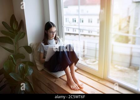 Cute teenager girl reading book at home while sitting on window sill Stock Photo