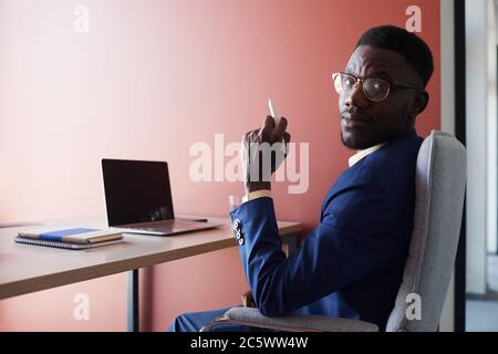 Portrait of elegant African-American man glancing at camera while using laptop at desk while working in contemporary office interior against red wall, Stock Photo