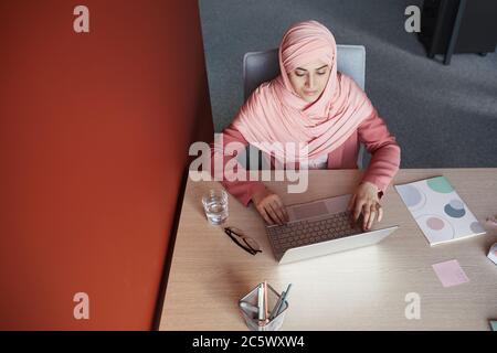 Top down view at young businesswoman wearing headscarf using laptop at desk while working in office, copy space Stock Photo