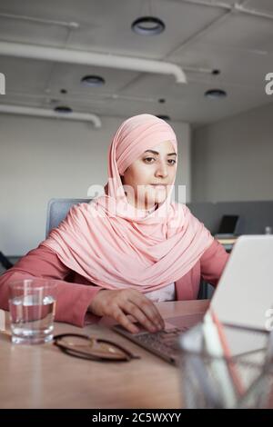 Vertical portrait of young businesswoman wearing headscarf using laptop at desk while working in office alone, copy space Stock Photo