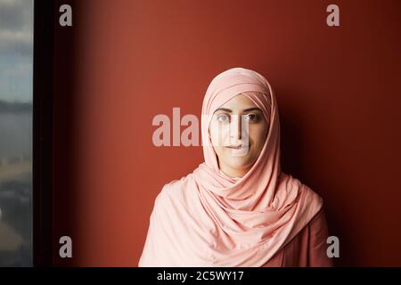 Head and shoulders portrait of contemporary young woman wearing headscarf looking at camera and smiling while standing against red wall, copy space Stock Photo
