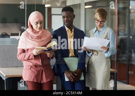 Waist up portrait of modern multi-ethnic business team of three holding documents while standing behind glass wall in office Stock Photo