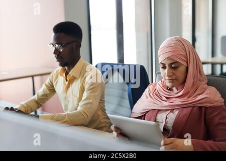 Portrait of young Muslim businesswoman using digital tablet while working in office with African-American man, copy space Stock Photo