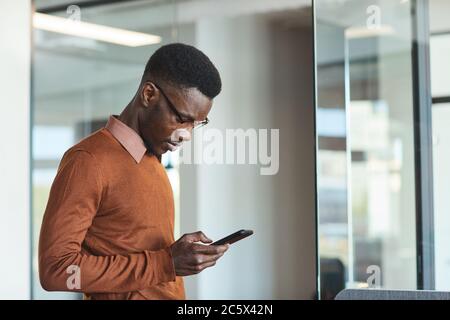 Side view portrait of trendy young African man using smartphone while standing in office or college interior, copy space Stock Photo