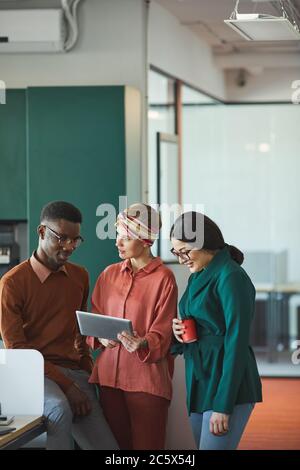 Vertical portrait of three contemporary business people discussing work in office, focus on young woman holding tablet and talking to colleagues, copy Stock Photo