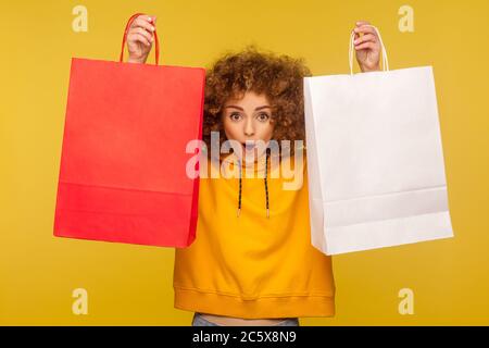 Portrait of shocked shopper, amazed curly-haired girl in hoodie raising shopping bags with blank area, copy space on packages for advertising text. in Stock Photo