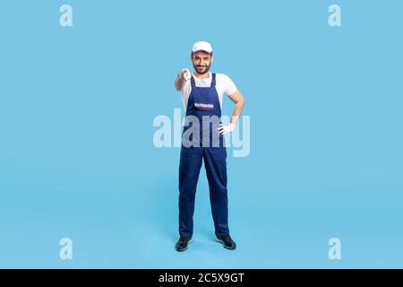 Full length happy courier in overalls, cap and protective gloves standing, pointing finger to camera, choosing you. Profession of service industry, lo Stock Photo